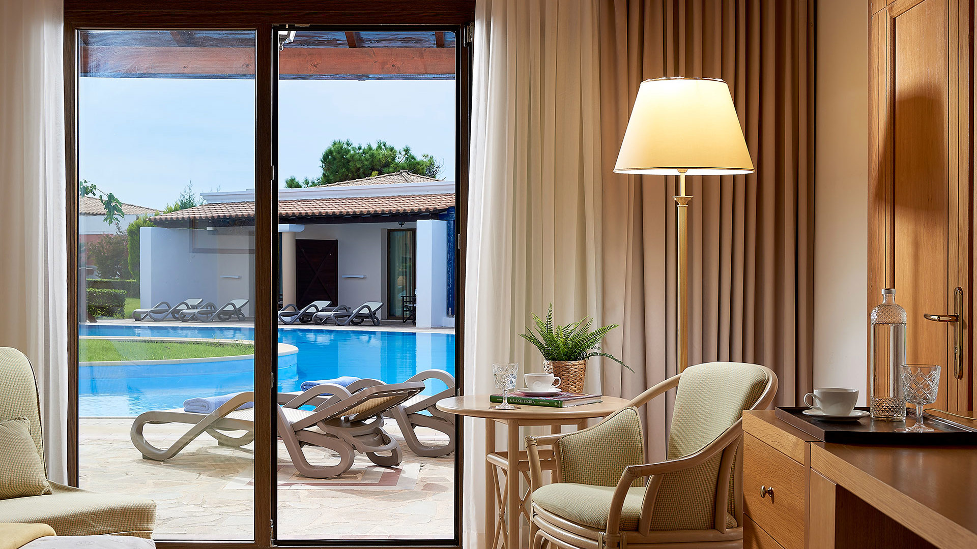 Room with an armchair, floor lamp and small table by glass doors opening to a terrace with sunbeds and a pool