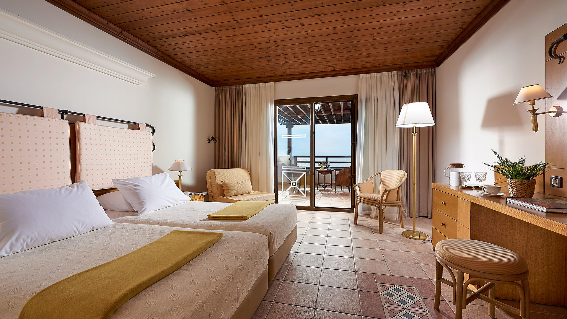 Room with a wooden ceiling, terracotta floor, two beds with mustard throws, armchairs and balcony doors with a sea view
