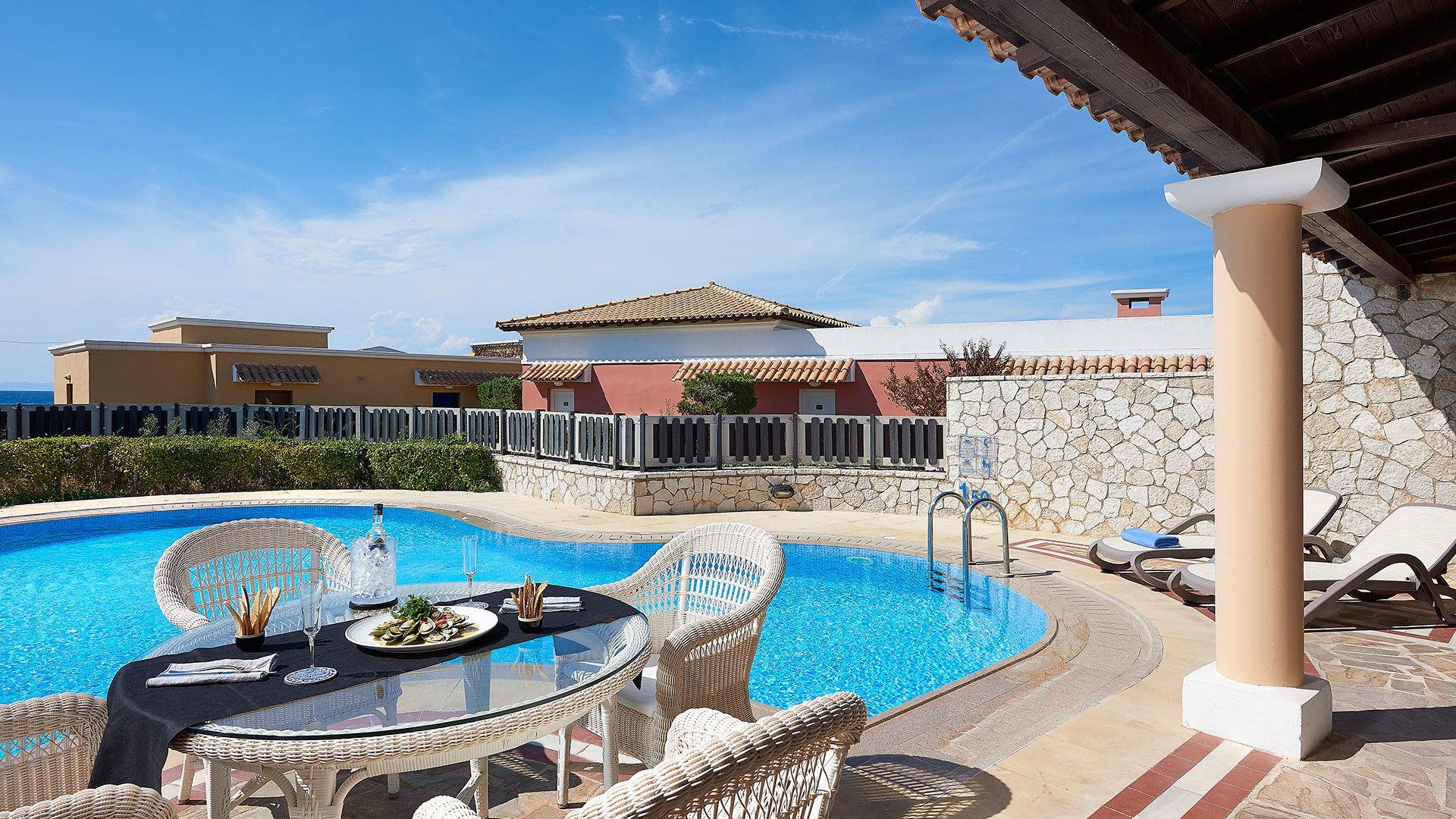Covered terrace with a column and a wicker dining table set with food beside a pool with sunbeds and a stone wall