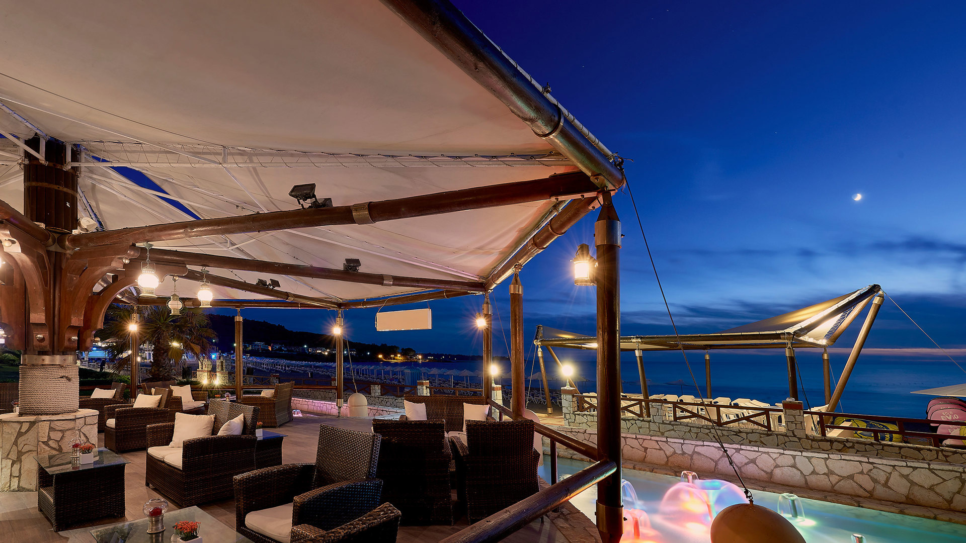 Lounge bar at night with sofas under a white canopy, glowing lights by the pool and the moon over the sea