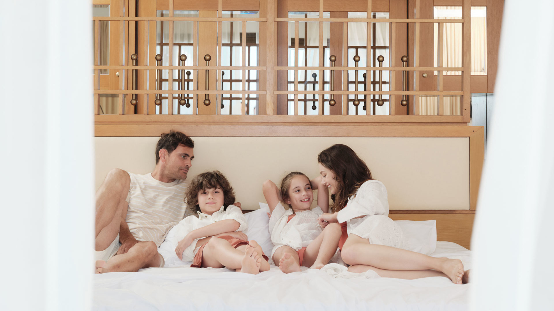 Family of four in white bathrobes relaxing together on a large bed, with a wooden mezzanine railing behind