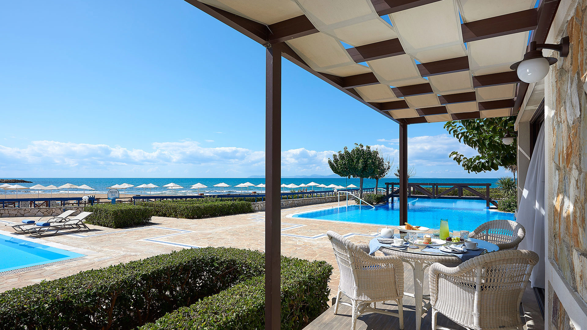 Covered terrace with a set wicker dining table beside pools with sunbeds, a green hedge and the sea beyond