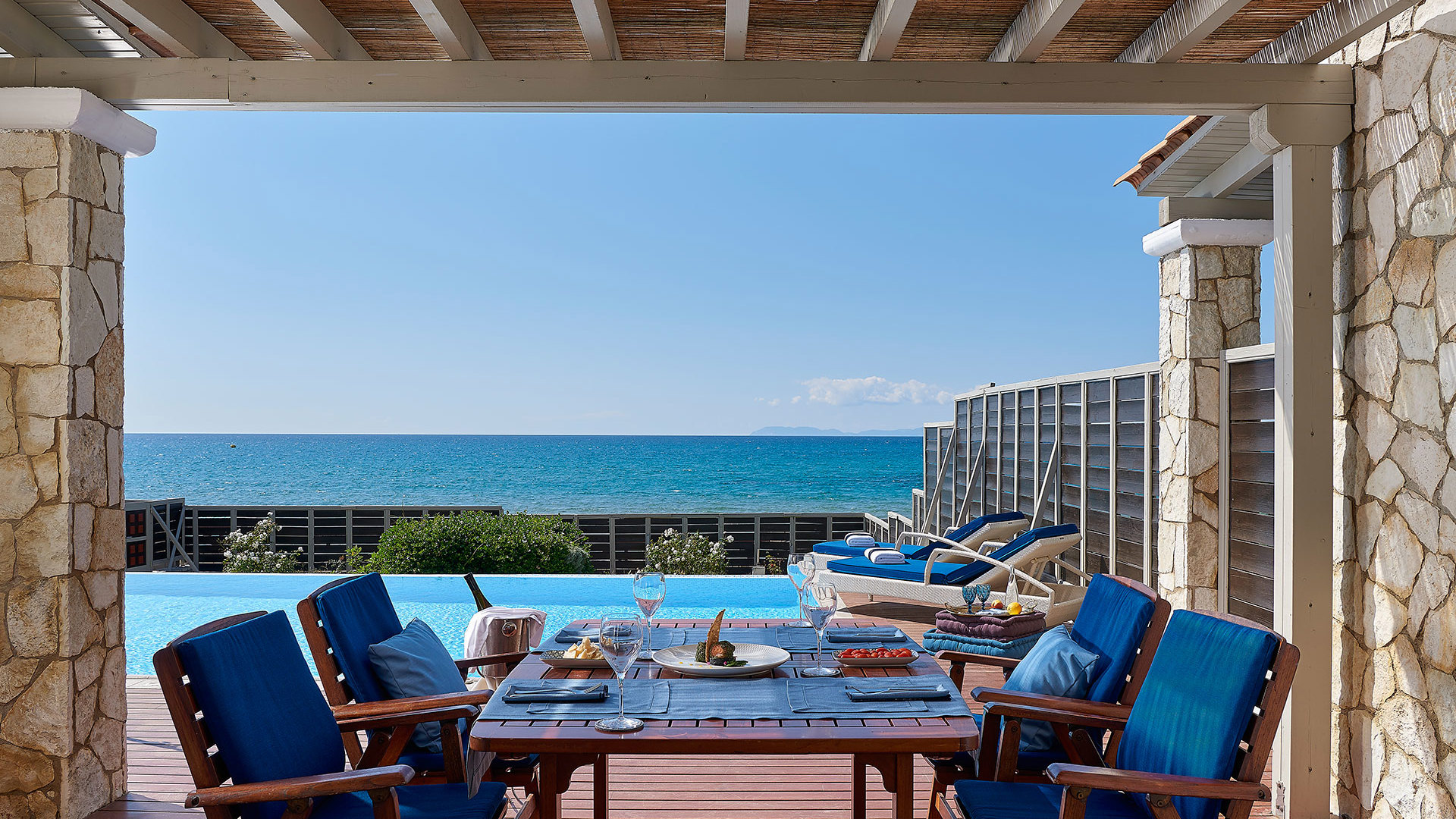 Covered terrace with stone columns, dining table with blue chairs, pool, sunbeds and sea view