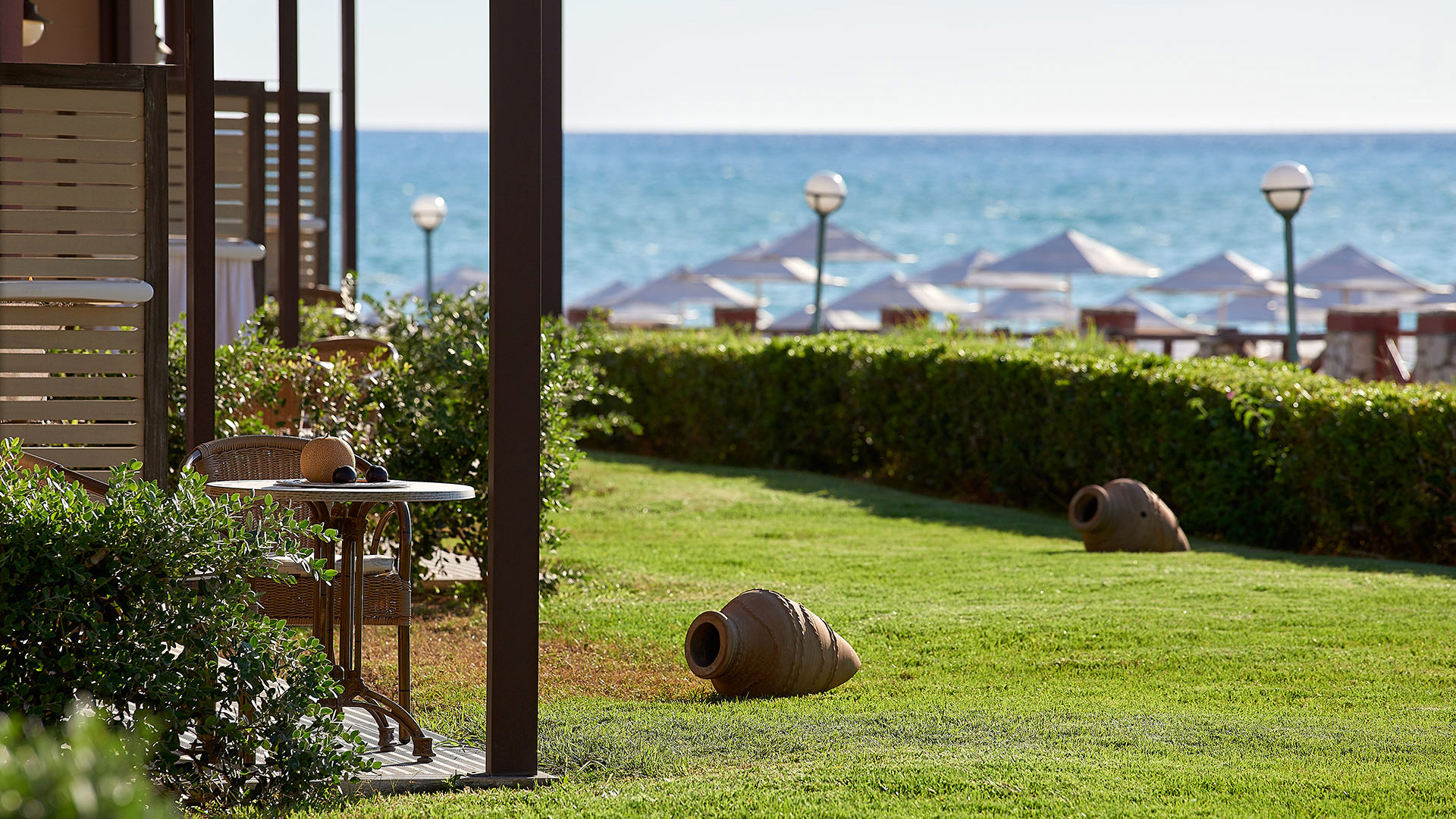 Terrace overlooking a green lawn with clay pots and a hedge, with beach umbrellas and the blue sea in the distance