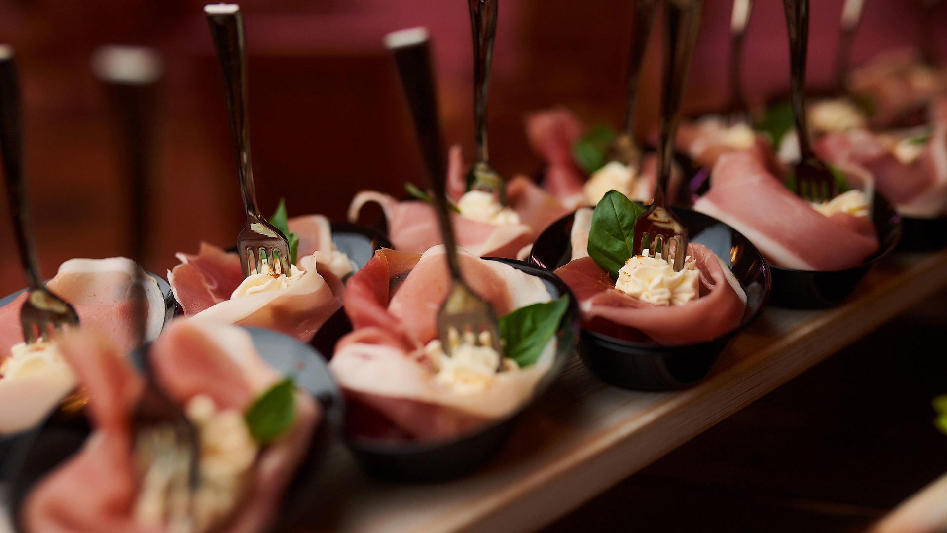 Bite-sized appetisers in black spoon bowls, with slices of prosciutto, soft cheese and fresh basil leaves