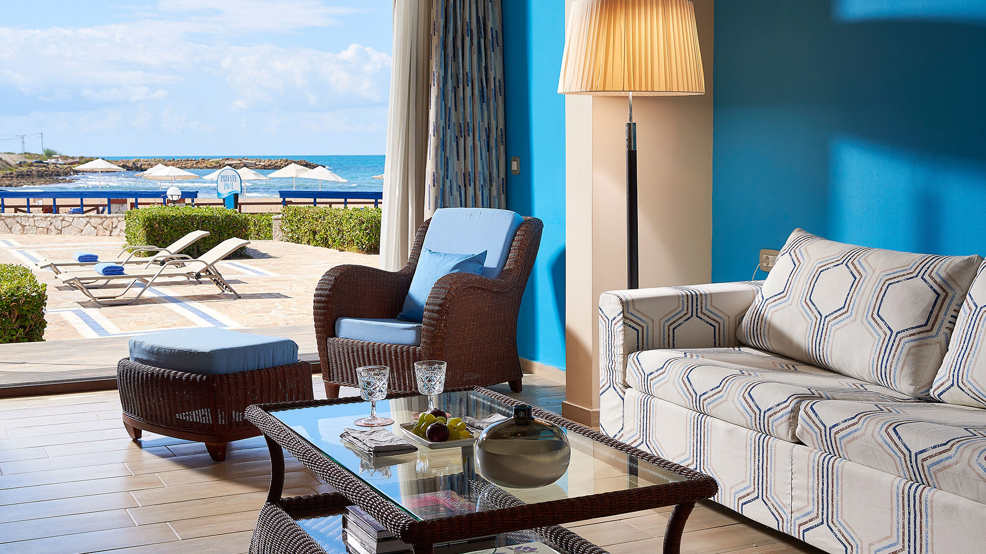 Lounge with a blue wall, striped sofa, wicker armchair, coffee table with glasses and open doors to sunbeds and the sea