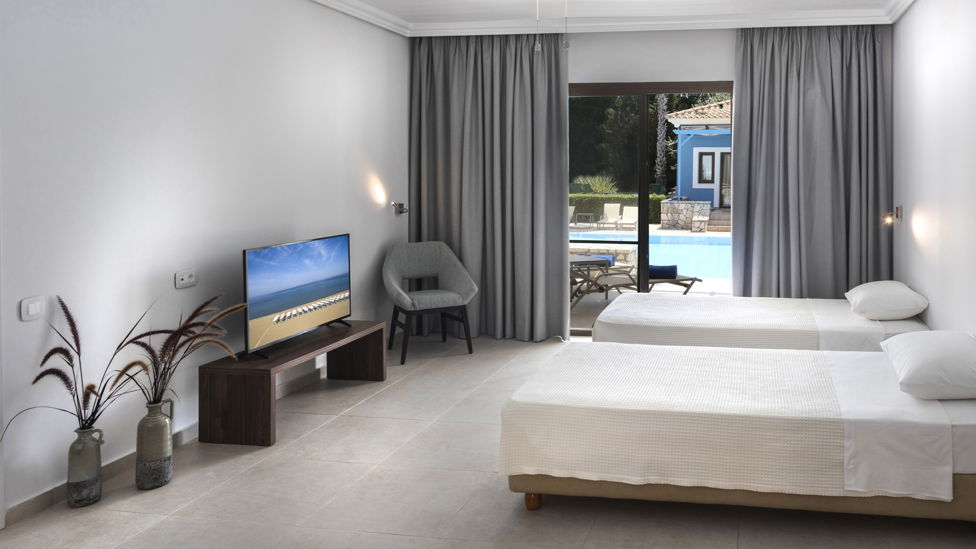 Room with two single beds, TV on a wooden stand, grey armchair, vases with dried grasses and glass doors to the pool