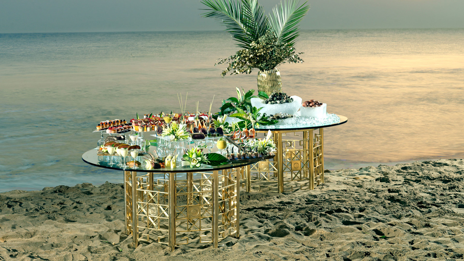 Canapés and finger food on glass tables with golden bases, set on the sandy beach as the sun sets over the sea