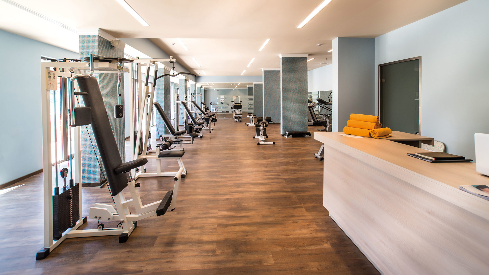Spacious fitness centre with rows of weight machines on a wooden floor and a reception desk with stacked orange towels