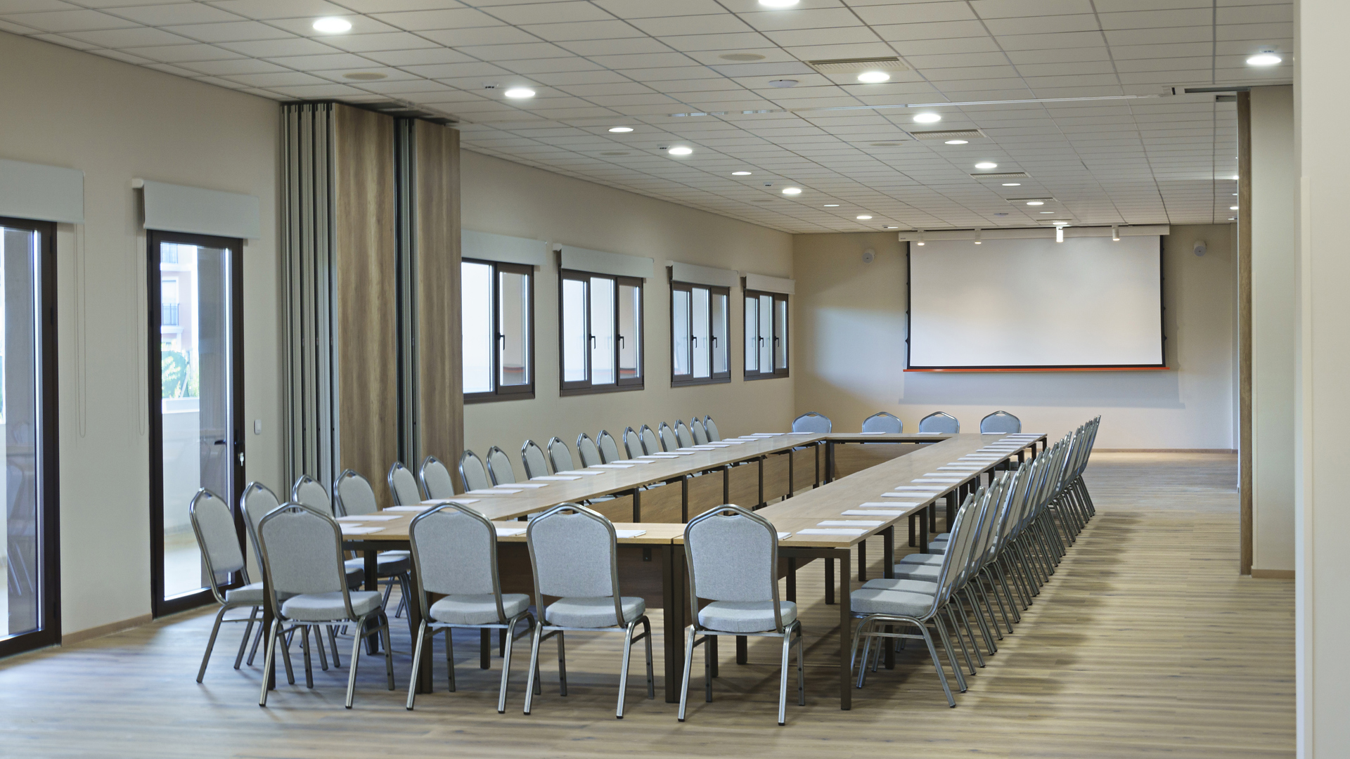 Modern meeting room with tables in a U-shape, grey chairs, a projection screen and a light wooden floor