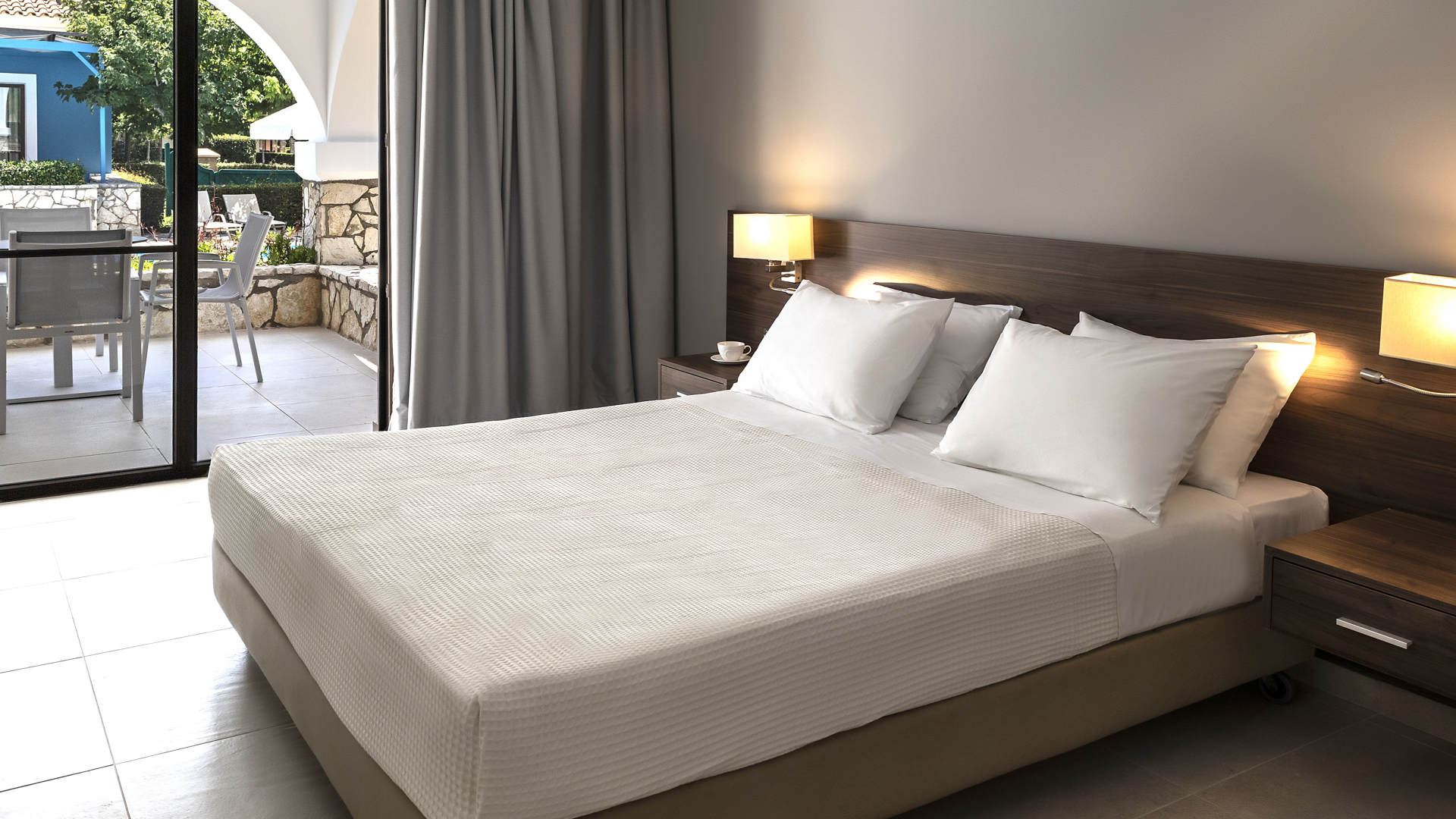 Double bed with white linen and wooden headboard with lamps, grey curtains and glass doors to a terrace with table and chairs
