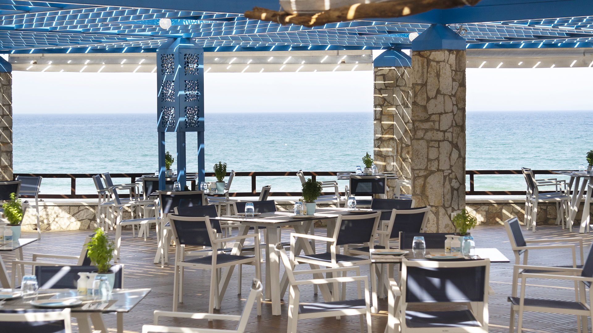 Seaside restaurant with blue pergola, stone columns, white chairs and potted herbs, facing the turquoise sea