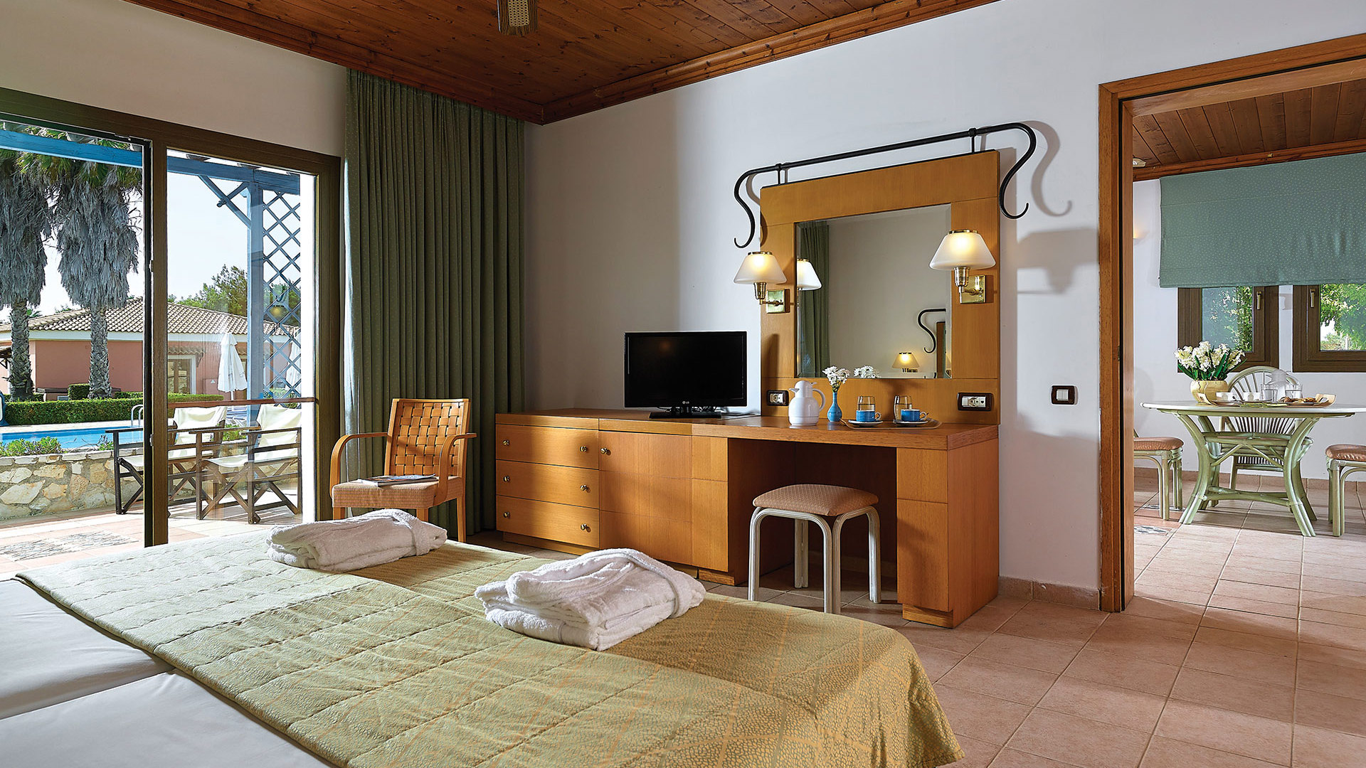 Room with wooden ceiling and fan, bed with towels, desk with TV and iron-framed mirror, and glass doors to a pool terrace