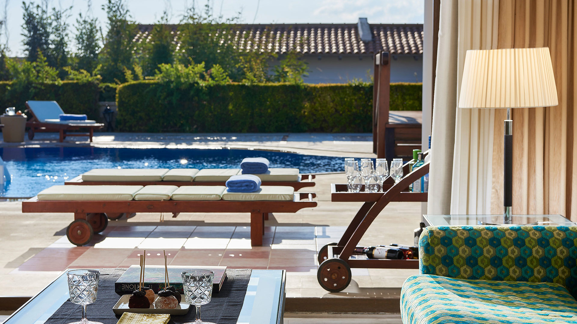 Patterned sofa, floor lamp and a table with glasses and snacks inside, opening to wooden sunbeds by a pool and greenery