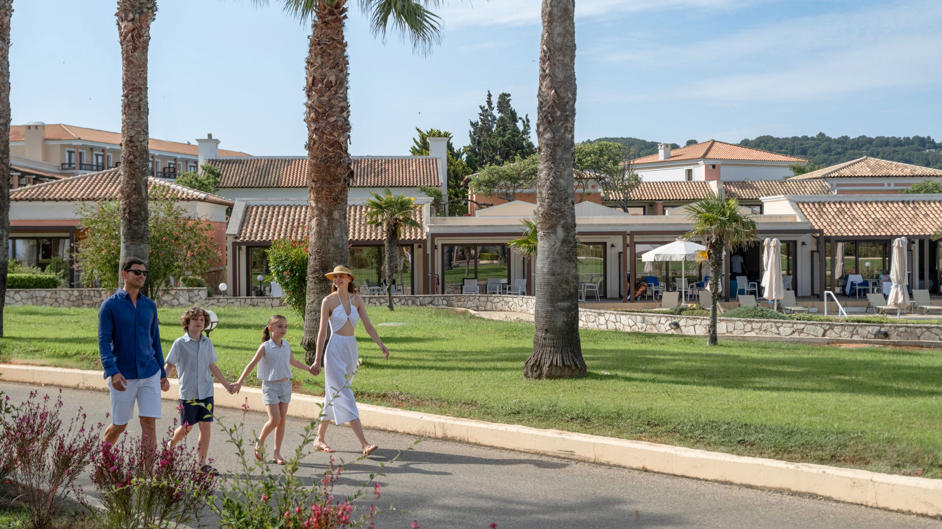Family with two children walking along a garden path lined with palm trees and flowers at Aldemar Olympian Village