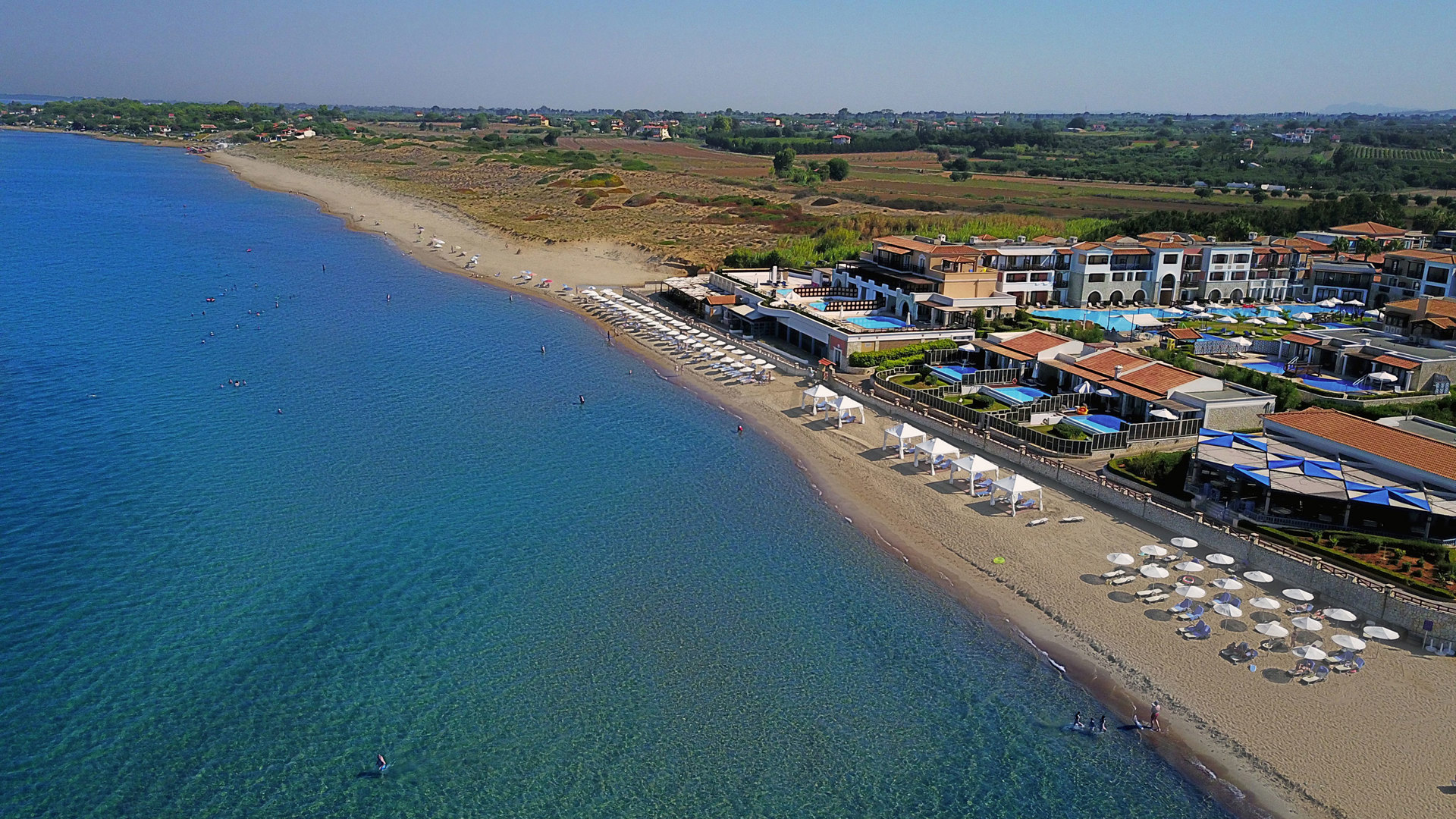 Aerial view of the long sandy beach of Skafidia, with umbrellas, pools and buildings set along the clear blue sea