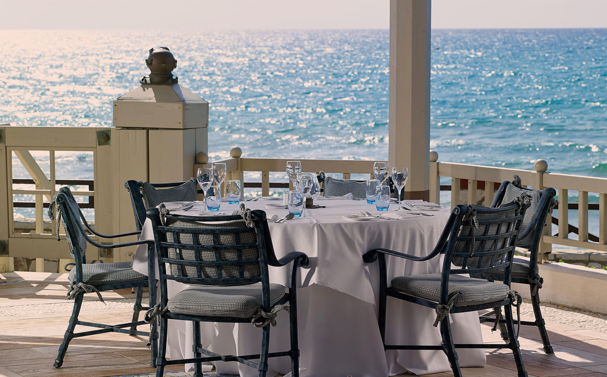 Round table with white linen and wrought iron chairs on a terrace, with the sunlit sea beyond the railing