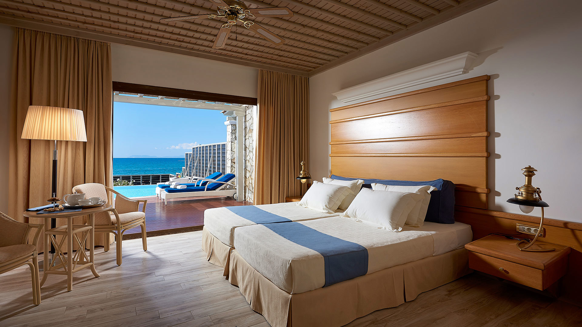 Bedroom with double bed, wooden ceiling and armchair, opening to a terrace with sunbeds, pool and sea view