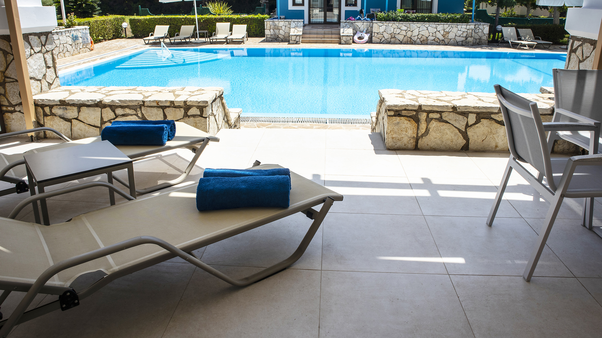 Shaded terrace with sunbeds and blue towels overlooking a pool with stone walls, white umbrellas and a blue building