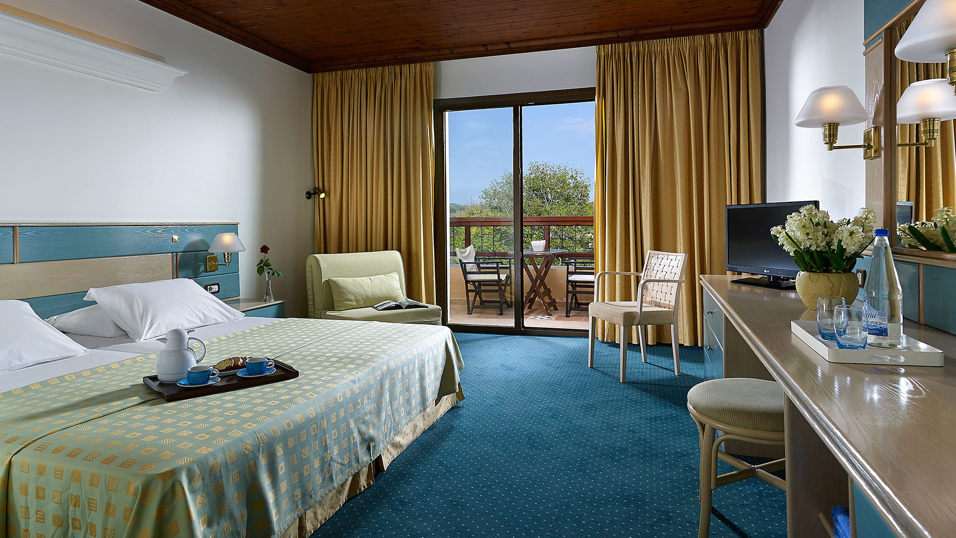 Room with a wooden ceiling, double bed, armchair, desk with mirror and flowers, and balcony doors framed by golden curtains