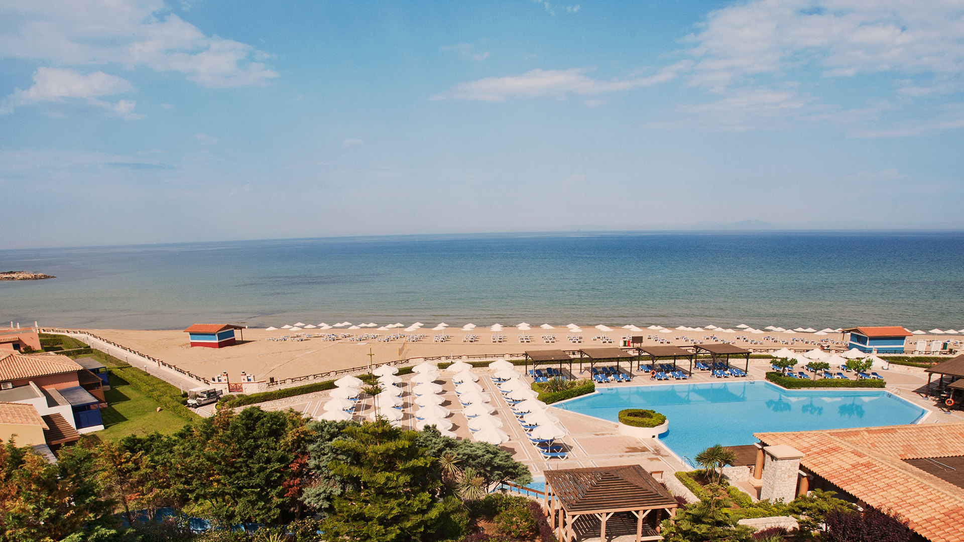 Panoramic view of the pool with sunbeds, the sandy beach and the Ionian Sea, framed by pine trees and tiled roofs