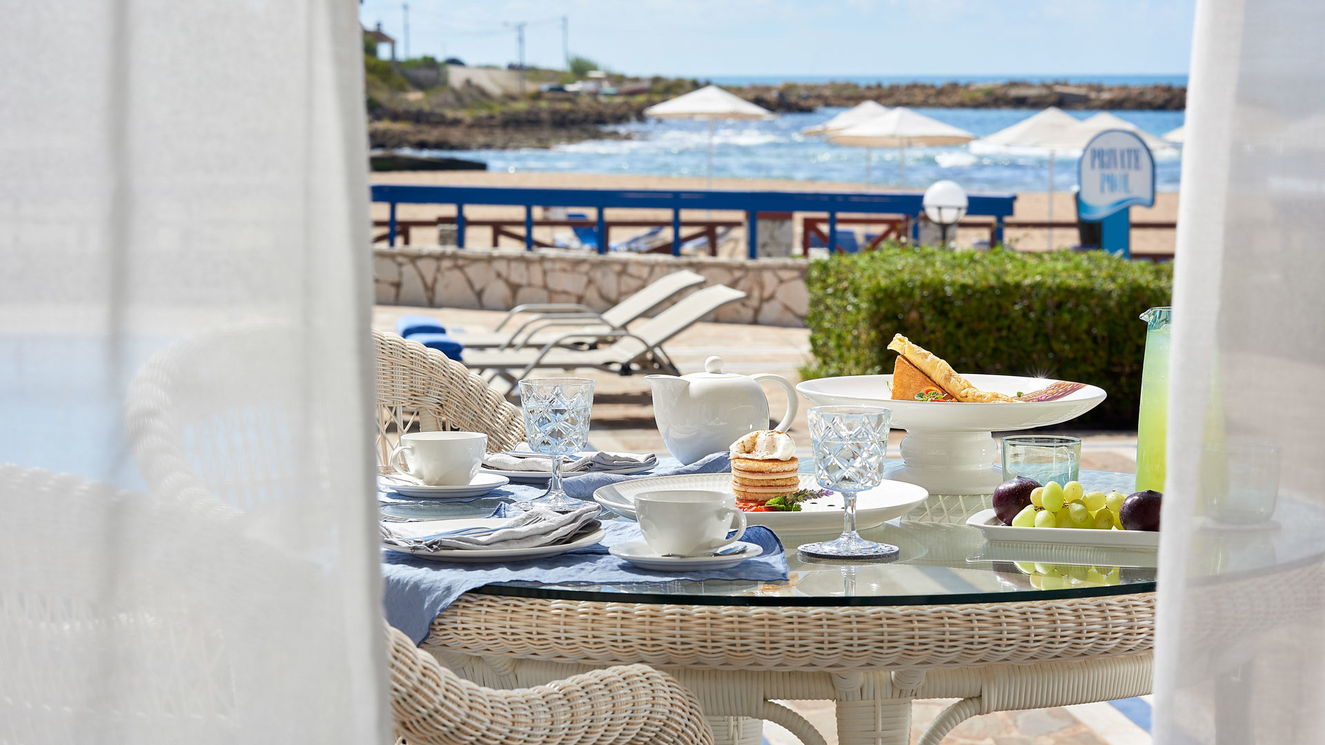 Breakfast with coffee, pastries and fruit on a terrace table, framed by white curtains, with sea view