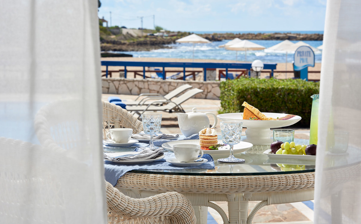 Breakfast with coffee, pastries and fruit on a terrace table, framed by white curtains, with sea view