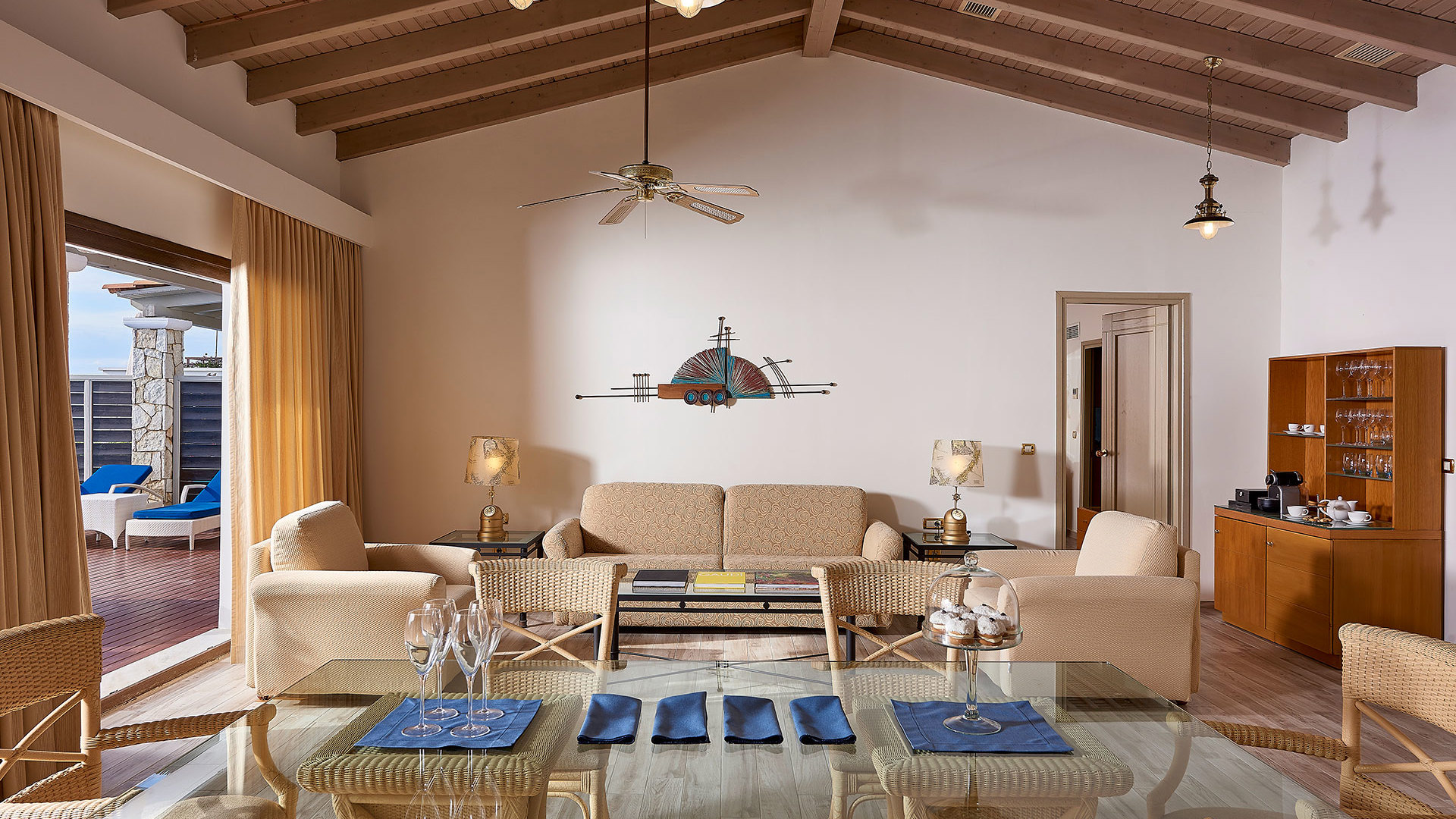 Living area with wooden beamed ceiling, sofa, armchairs, glass dining table and terrace doors opening to the pool