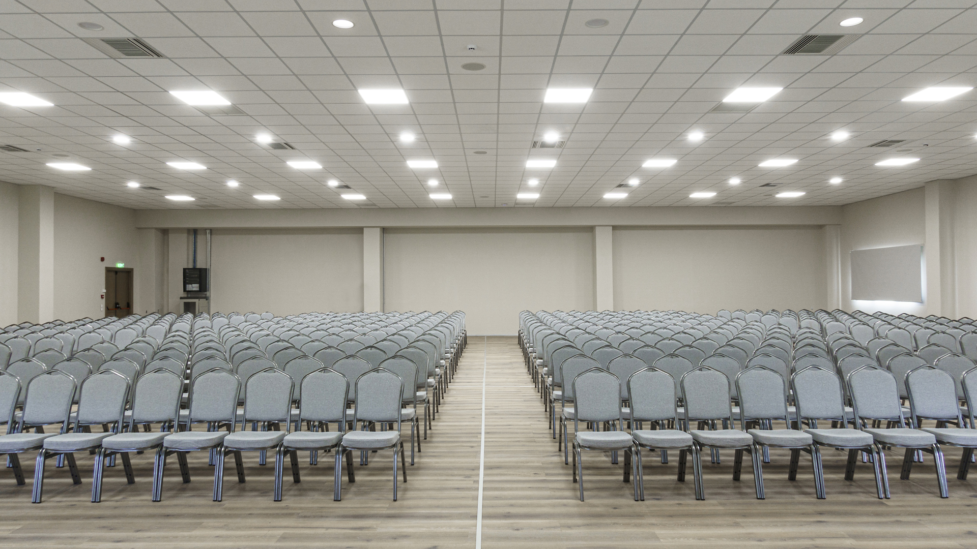 Bright hall with rows of grey chairs on either side of a central aisle, a wooden floor and panel lights above