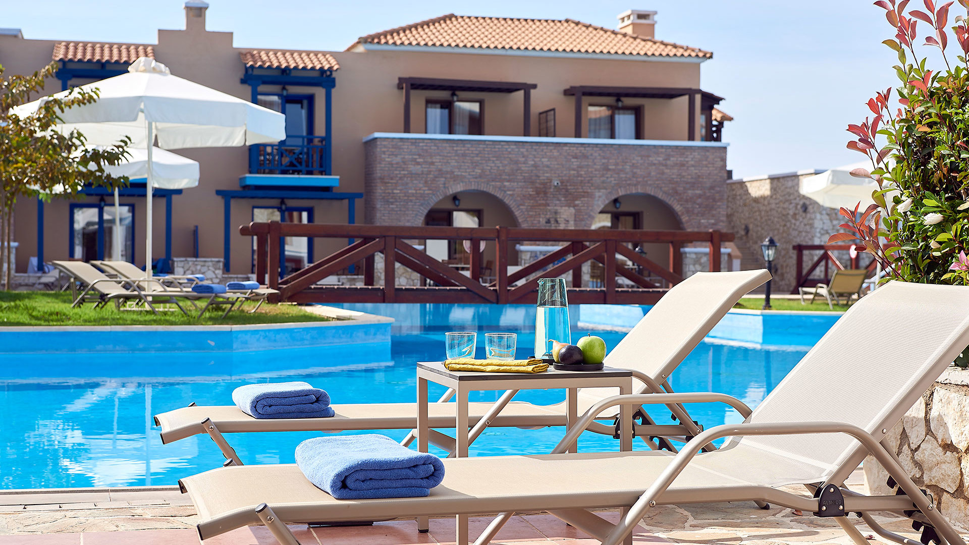 Two sunbeds with blue towels and drinks by a pool with a wooden bridge, in front of a stone building with balconies