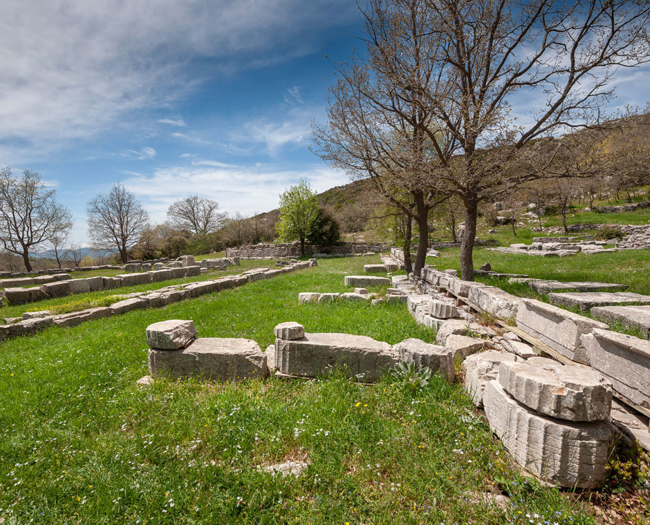 Ancient column drums and stone ruins on green grass at the archaeological site of Olympia, under a blue sky