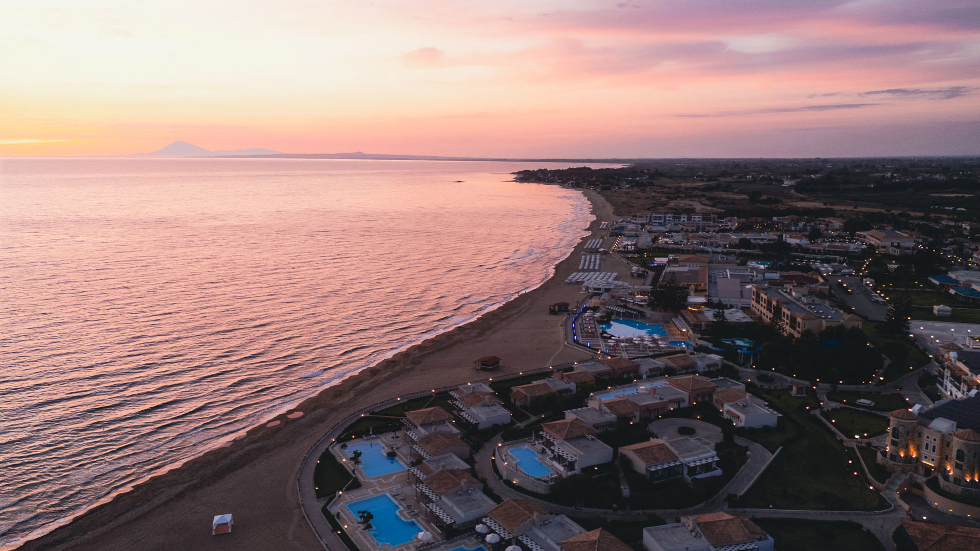 Aerial view of Aldemar Olympian Village at sunset, with lit pools along Skafidia beach and the Ionian Sea in pink hues