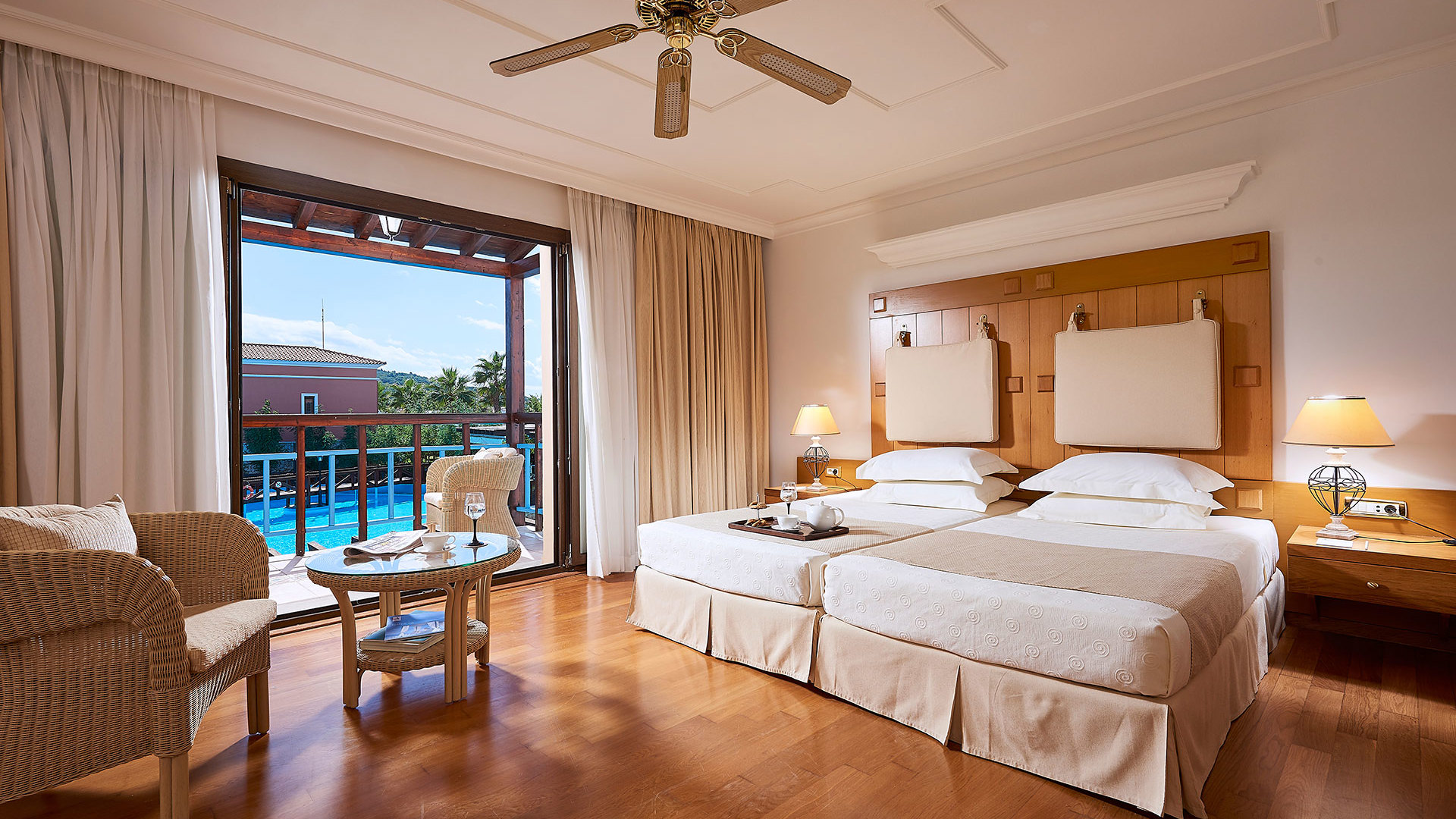Room with wooden floor, two white beds, ceiling fan, wicker armchairs and balcony doors overlooking a pool and palm trees