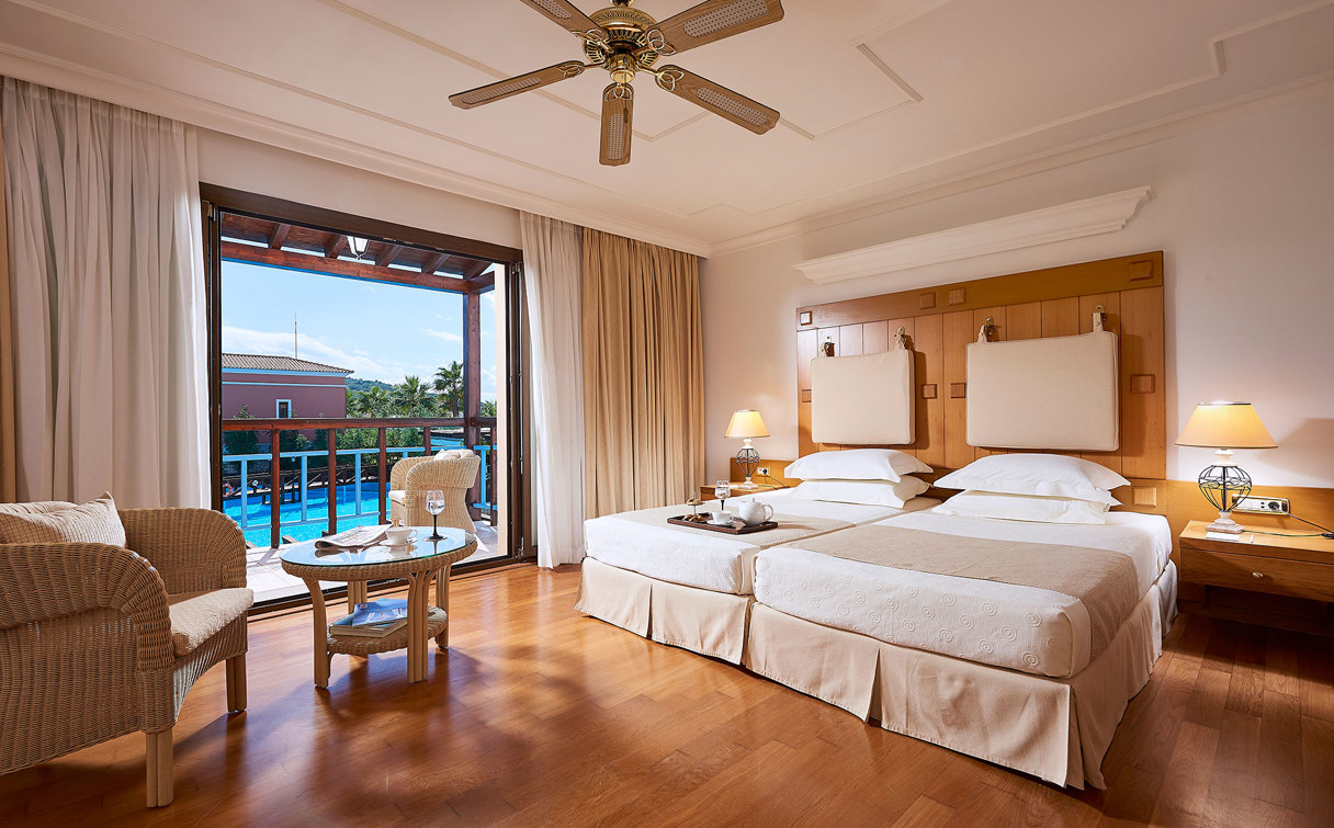 Room with wooden floor, two white beds, ceiling fan, wicker armchairs and balcony doors overlooking a pool and palm trees