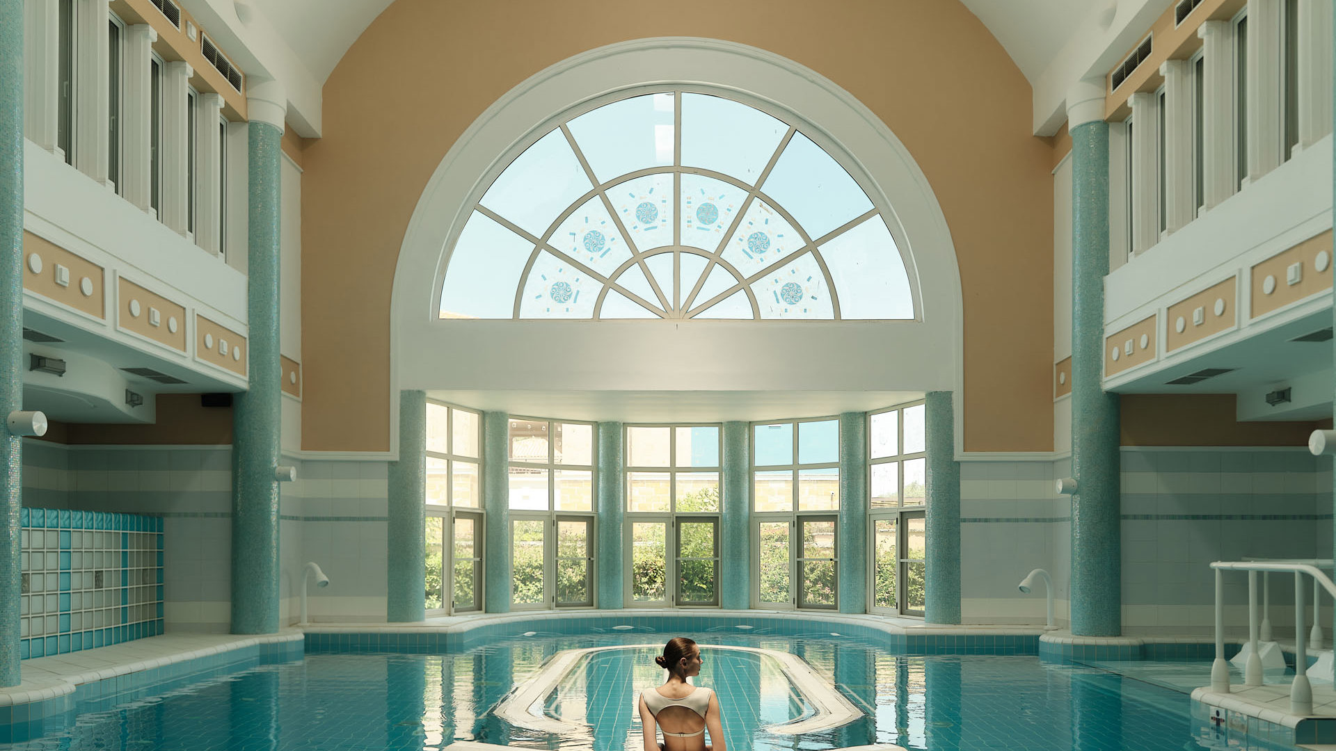 Woman seen from behind sitting at the center of an indoor pool, facing a tall arched window flooded with daylight