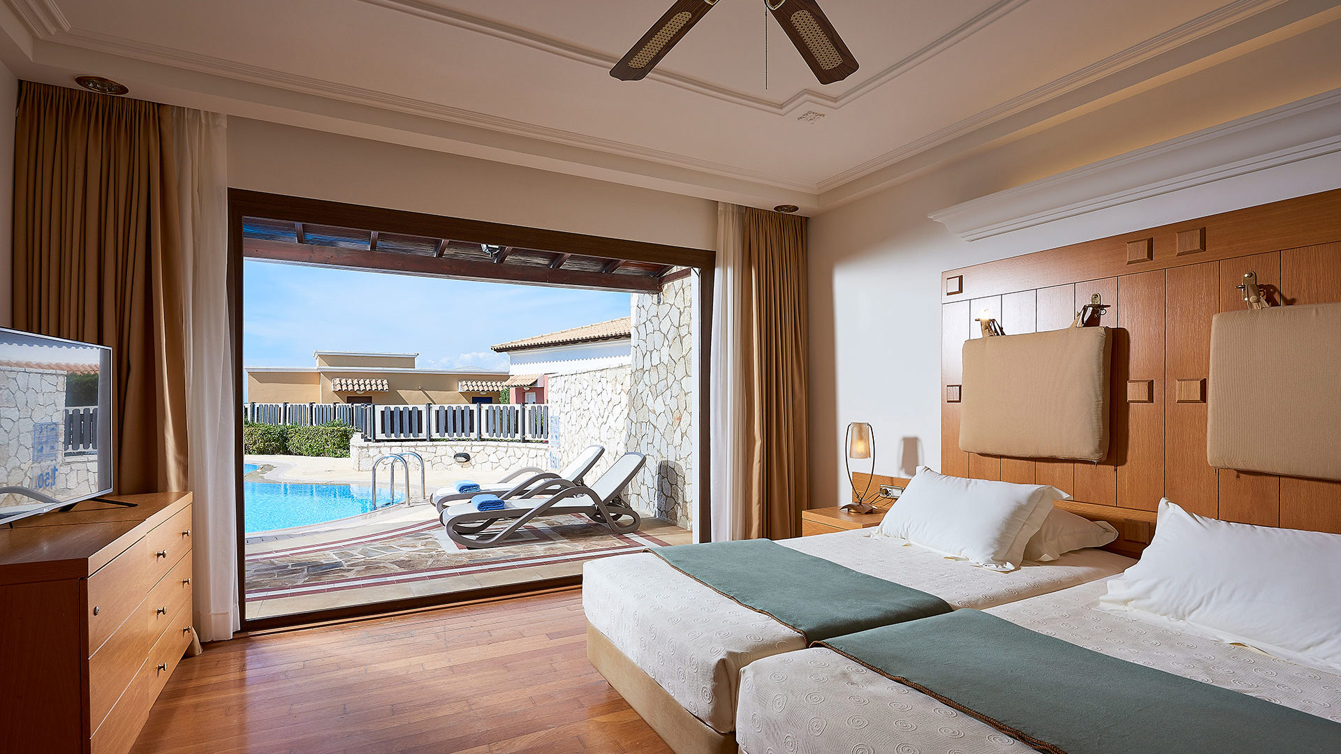 Bedroom with two beds, wooden headboard wall, ceiling fan, wooden floor and open doors to a terrace with sunbeds and a pool