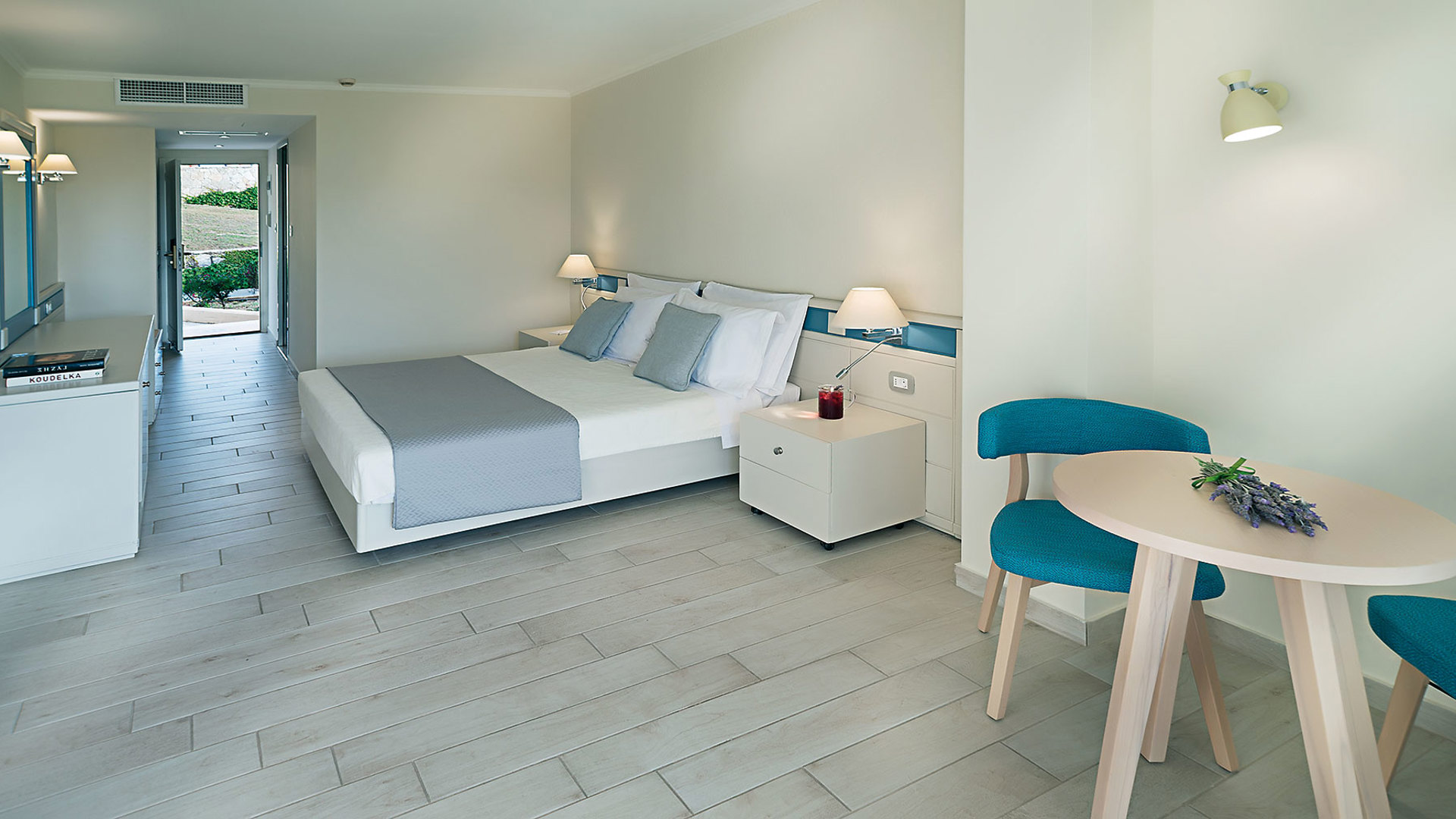 Bright room with a double bed, light floor tiles, a round table with two teal chairs and an open door to the garden