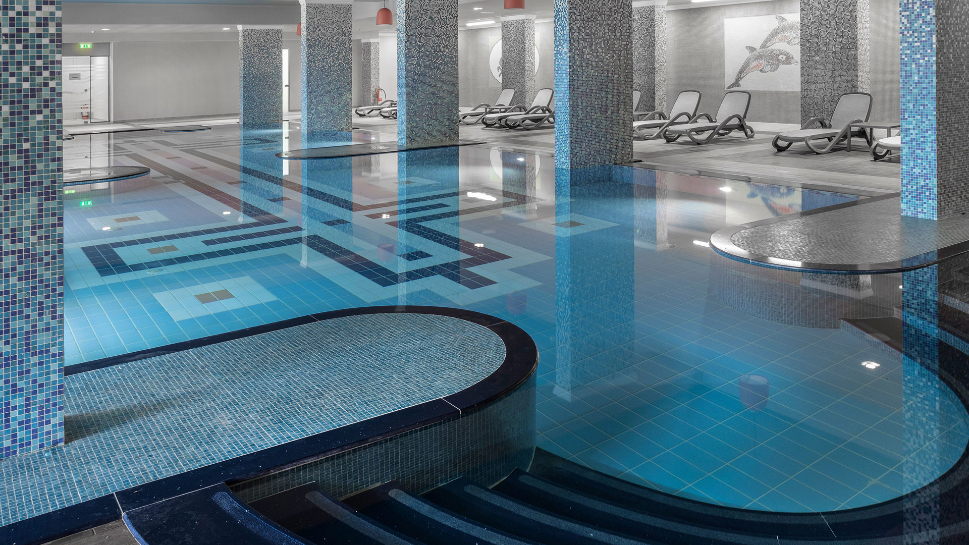 Curved tiled steps leading into an indoor pool with mosaic columns, sunbeds and a dolphin mosaic on the wall