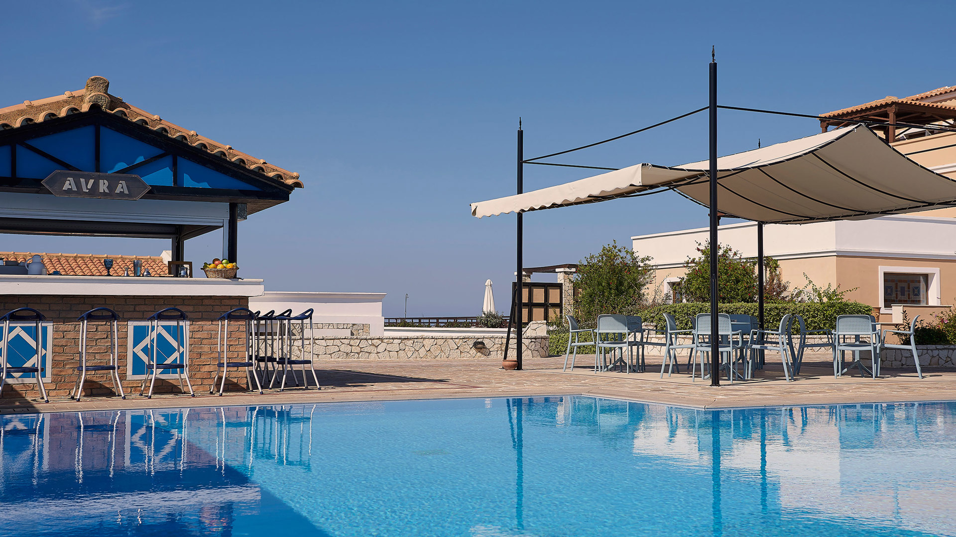 Aura pool bar with tiled roof and stools beside the pool, and tables shaded by a white sail under a clear blue sky