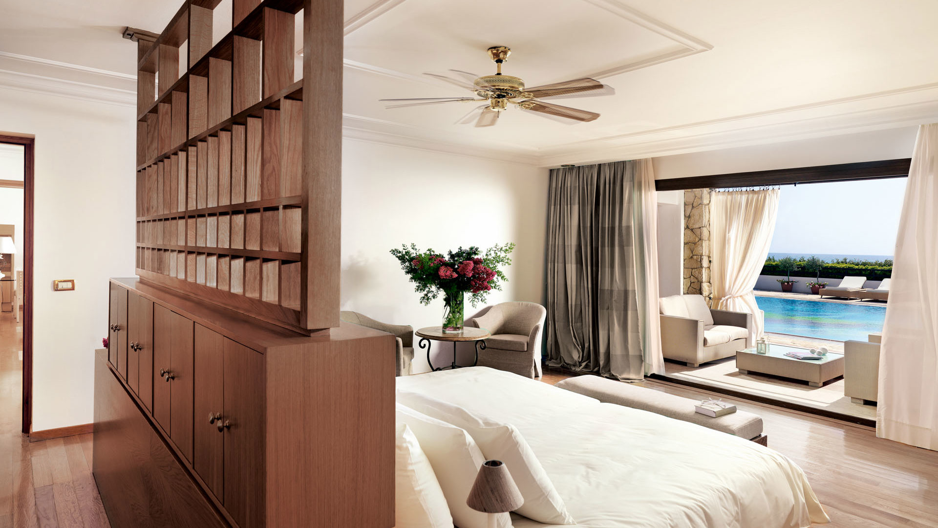 Bright bedroom with wooden shelves, double bed and ceiling fan, opening to a terrace with armchairs and pool view
