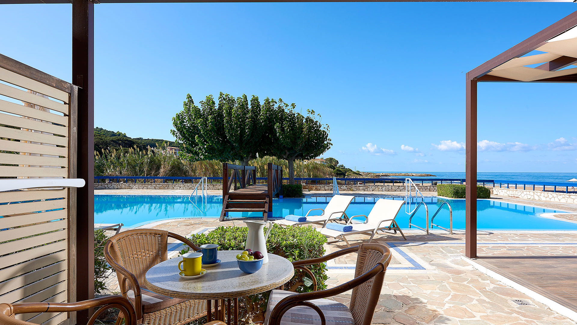 Shaded terrace with wicker chairs and a round table overlooking a pool with sunbeds, trees and the sea in the distance