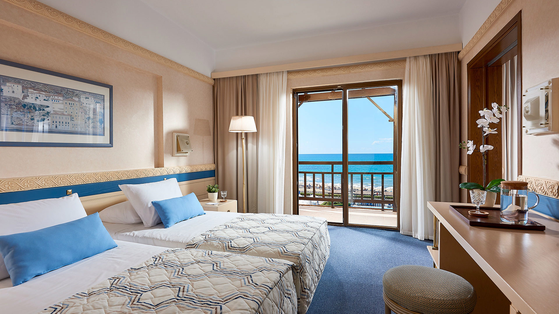 Room with two beds and blue cushions, artwork above, desk with an orchid, blue carpet and balcony door with a sea view