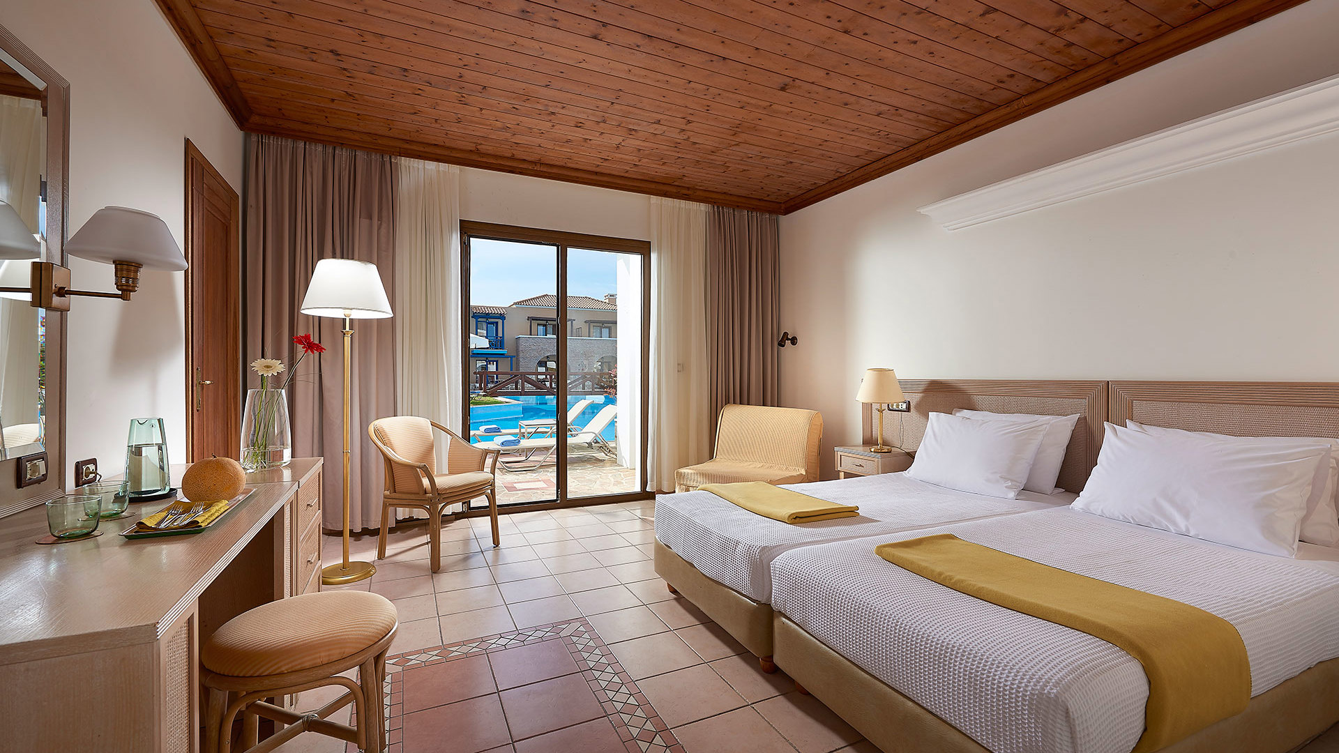 Room with a wooden ceiling, terracotta floor, two beds with mustard throws, desk, armchair and glass door with pool view