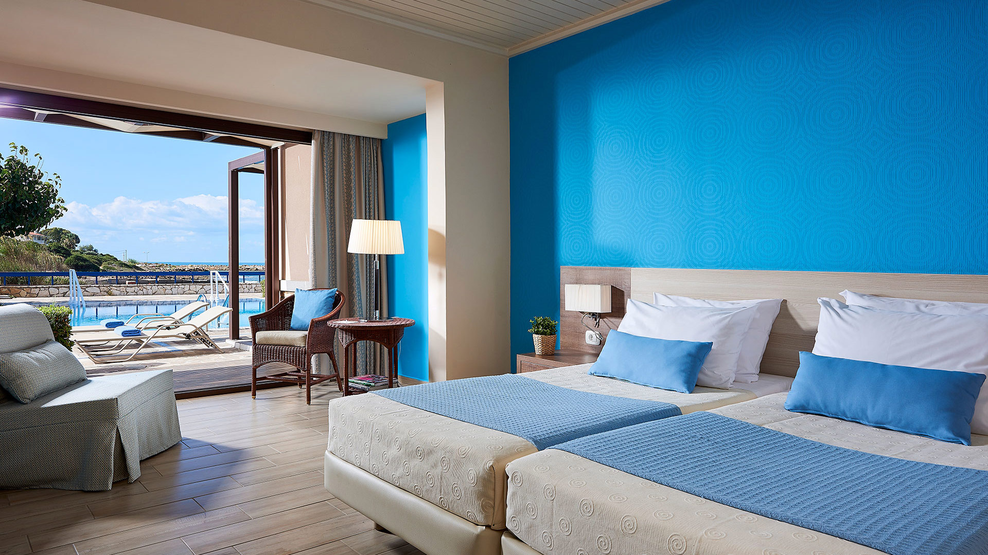 Room with a bright blue wall, two beds with blue throws, armchairs and open doors to a terrace with a pool and sea view
