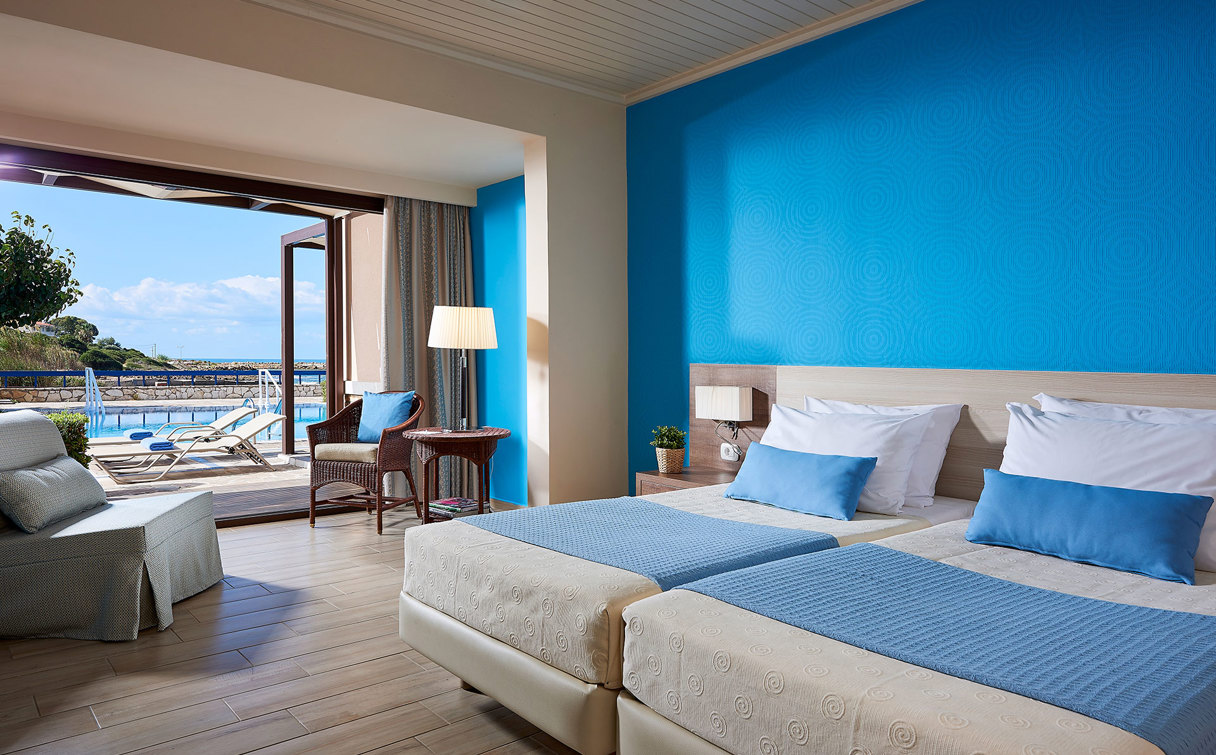 Room with a bright blue wall, two beds with blue throws, armchairs and open doors to a terrace with a pool and sea view