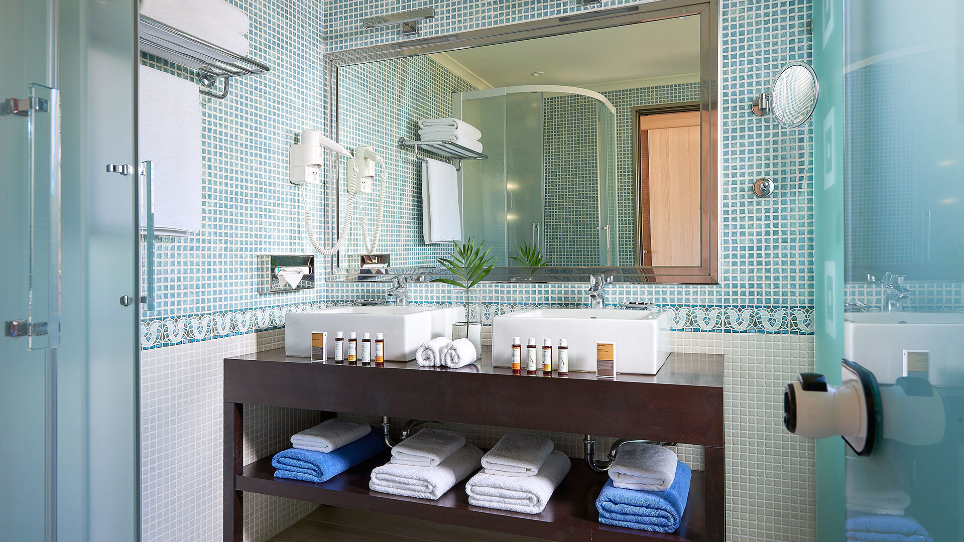 Bathroom with turquoise mosaic tiles, mirror, two white basins and a dark wooden vanity with folded white and blue towels