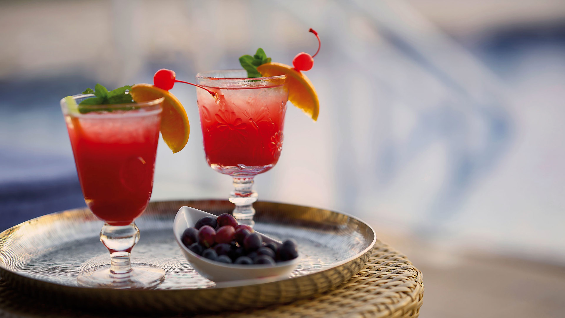 Two red cocktails with orange slices, cherries and mint on a tray with berries, beside the pool