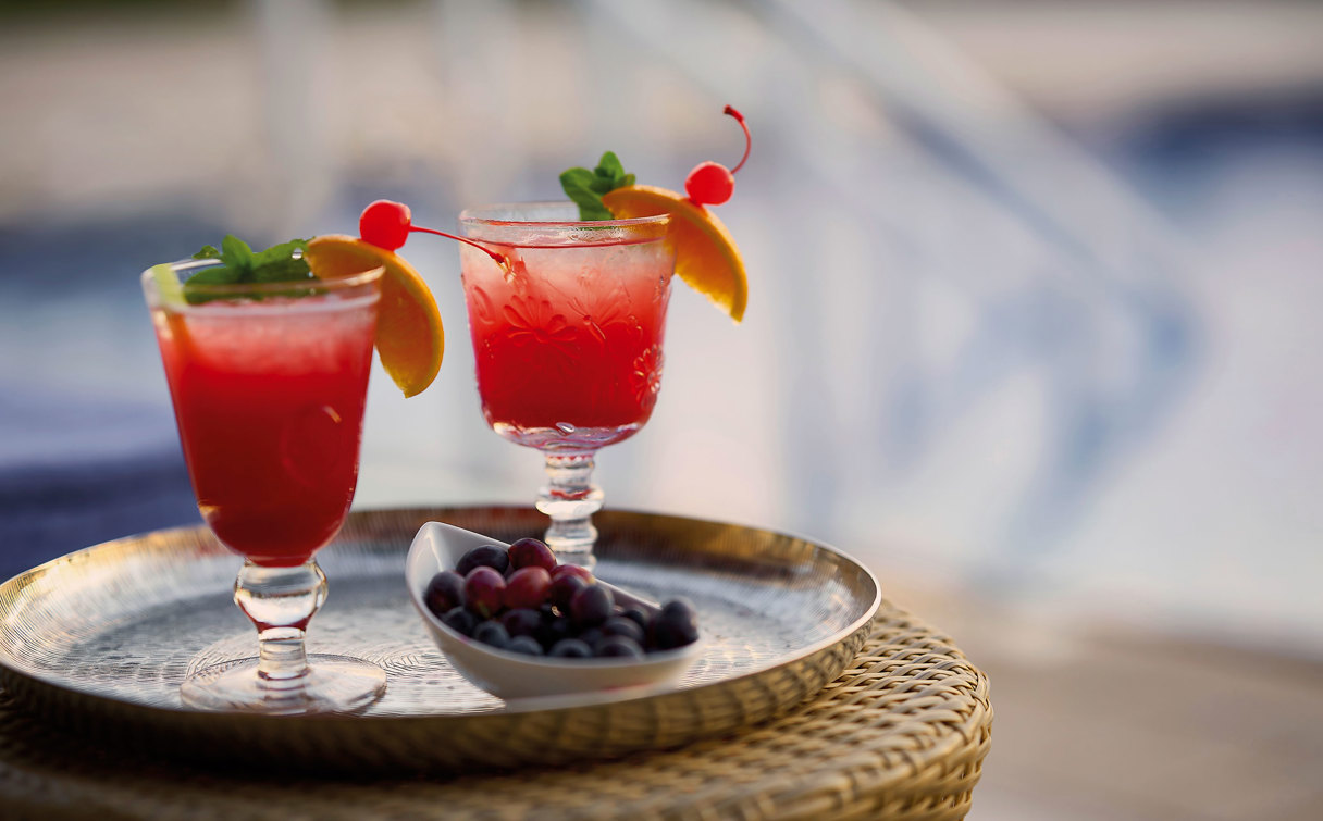 Two red cocktails with orange slices, cherries and mint on a tray with berries, beside the pool