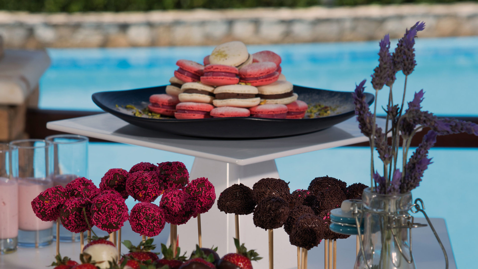 Dessert table with macarons, colourful cake pops and chocolate-dipped strawberries, with the pool and beach behind