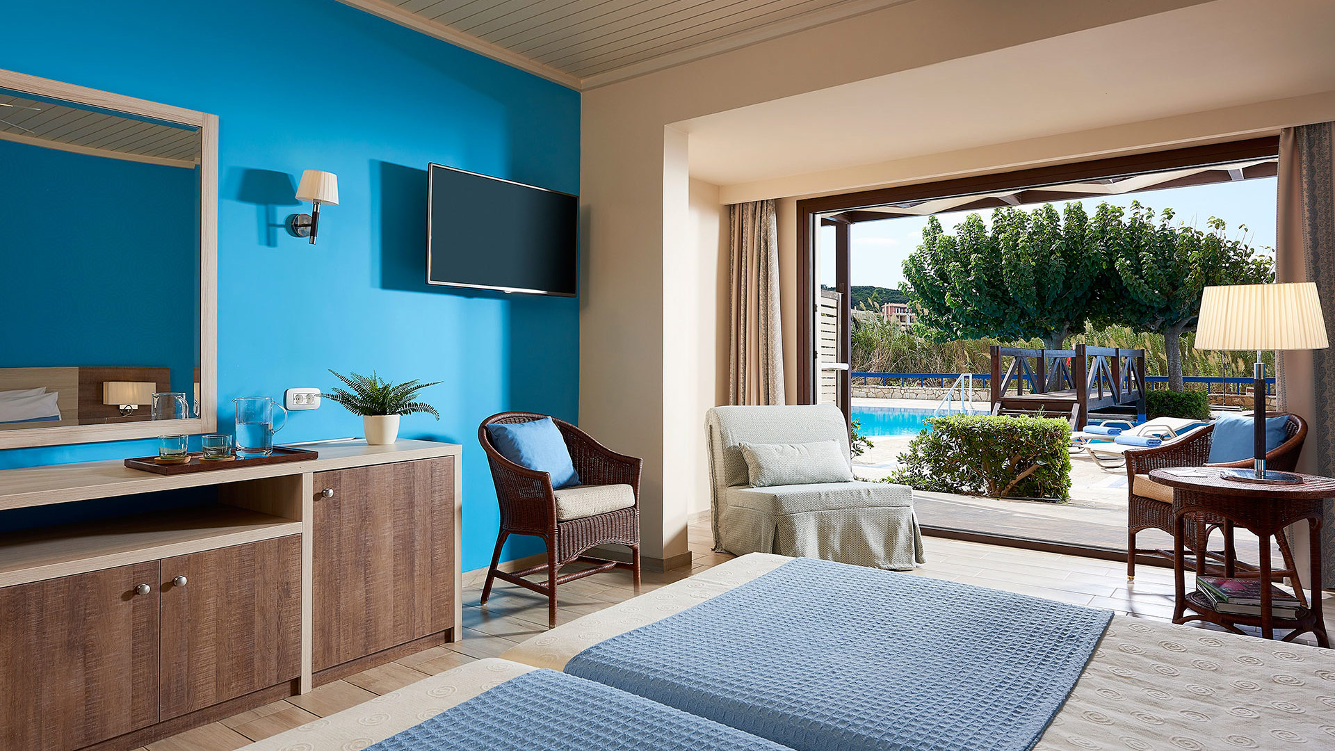 Room with a bright blue wall, TV, wooden dresser, wicker armchair and open doors to a terrace by the pool with trees